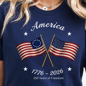 May include: Navy blue t-shirt with the word "America" in white script above two crossed American flags. The flags are red, white, and blue. The text "1776 - 2026" and "250 Years of Freedom" are below the flags.