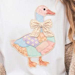 May include: White t-shirt featuring a patchwork goose design. The goose has a pink head, a red beak, and a bow tie. The body is made of various patterned fabrics in pastel colors. The design is outlined with small white beads.