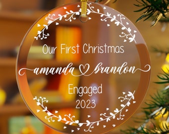 May include: A clear glass Christmas ornament with a white floral wreath design. The ornament says "Our First Christmas amanda ♥ brandon Engaged 2023".