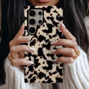 May include: A phone case featuring a cream base with a pattern of black, irregular shapes. The case has a camera module with three lenses. The phone case is held by a person wearing a white sweater.