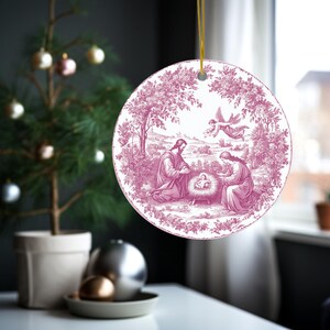 May include: A round Christmas ornament with a pink and white illustration of the Nativity scene. The ornament has a gold string for hanging and features a detailed depiction of Mary, Joseph, and baby Jesus.