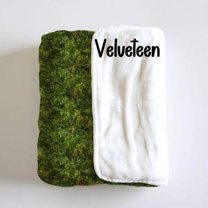 May include: A white, plush blanket with a green moss pattern on one side. The blanket has the word "Velveteen" printed on the white side.