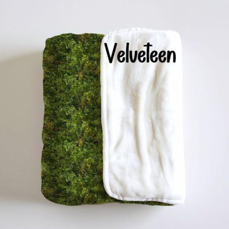 May include: A white, plush blanket with a green moss pattern on one side. The blanket has the word "Velveteen" printed on the white side.