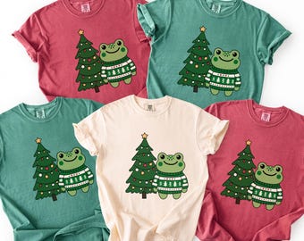 May include: A collection of short-sleeved t-shirts in various colors, each featuring a cartoon frog wearing a sweater and standing next to a decorated Christmas tree. The frog and tree are in a whimsical style.