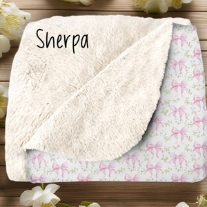 May include: A white sherpa blanket with a pink and white bow pattern on one side. The blanket has the word "Sherpa" written on the white side.