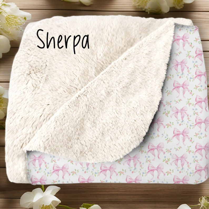 May include: A white sherpa blanket with a pink and white bow pattern on one side. The blanket has the word "Sherpa" written on the white side.