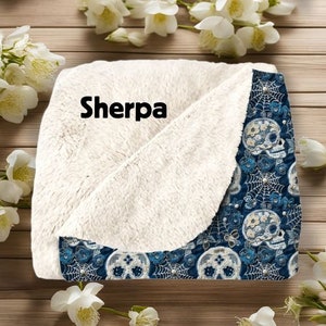 May include: A blue and white sherpa blanket with a floral skull pattern. The blanket has a soft, fluffy sherpa lining and a printed cotton outer layer. The text "Sherpa" is printed on the blanket.