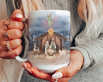 May include: White ceramic mug with a Christmas nativity scene design. The scene features a stable with a manger, a donkey, a sheep, Mary, Joseph, and a baby Jesus. An angel is above the stable with a star.