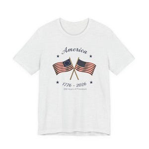 May include: A light gray t-shirt featuring a graphic with two crossed American flags. The flags are red, white, and blue, with the text "America" above and "1776 - 2026 250 Years of Freedom" below the flags.