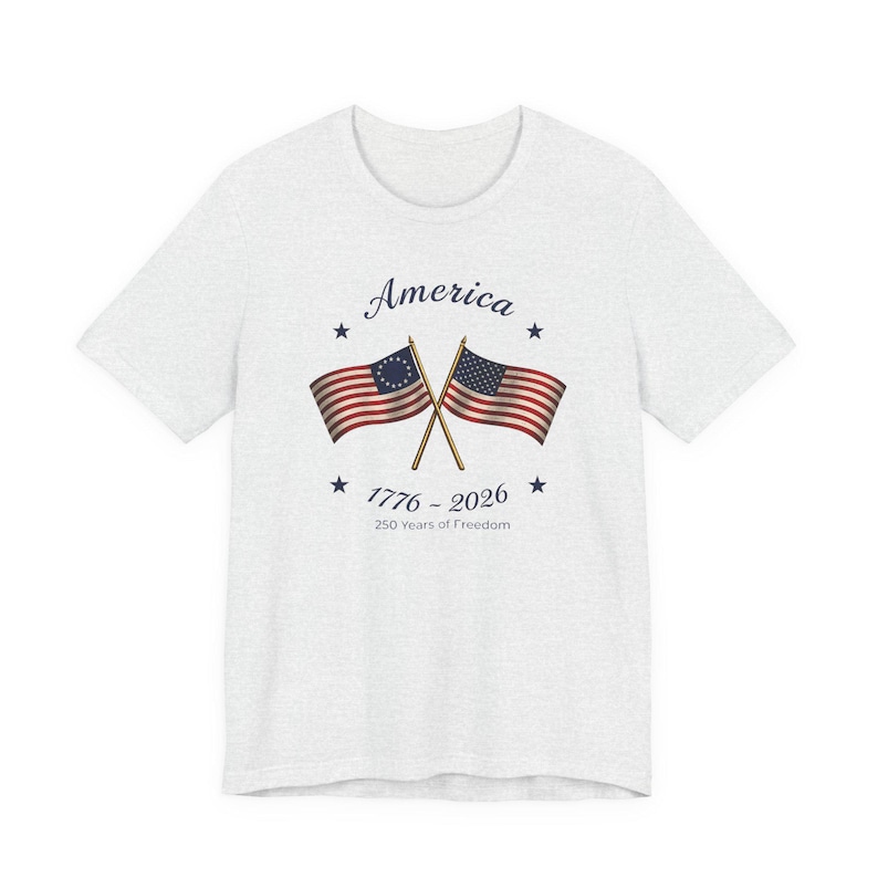 May include: A light gray t-shirt featuring a graphic with two crossed American flags. The flags are red, white, and blue, with the text "America" above and "1776 - 2026 250 Years of Freedom" below the flags.