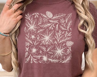 May include: A maroon t-shirt featuring a white floral design. The design includes poinsettias, pinecones, and various leaves and berries arranged in a square pattern. The shirt is made of a soft material.