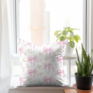 K&ouml;nnte beinhalten: Ein wei&szlig;es Kissen mit einem Muster aus rosa Schleifen. Die Schleifen sind mit Aquarellfarben bemalt und haben eine hellrosa Farbe. Der Hintergrund ist wei&szlig; mit kleinen blauen Blumen.