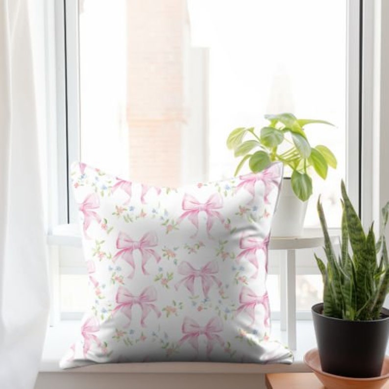 K&ouml;nnte beinhalten: Ein wei&szlig;es Kissen mit einem Muster aus rosa Schleifen. Die Schleifen sind mit Aquarellfarben bemalt und haben eine hellrosa Farbe. Der Hintergrund ist wei&szlig; mit kleinen blauen Blumen.