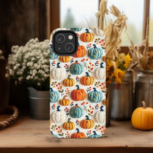Peut inclure: Une coque de t&eacute;l&eacute;phone blanche avec un motif de citrouilles orange, blanches et bleues. La coque a un d&eacute;coupage pour l'objectif de l'appareil photo noir.