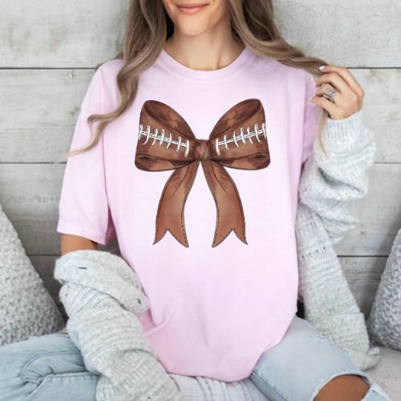 May include: A pink t-shirt with a brown football-shaped bow on the front. The bow has white stitching details.