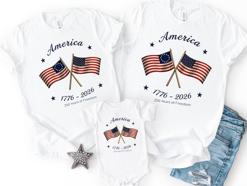 May include: White t-shirts and a baby onesie with a patriotic design. The shirts feature the word "America" above crossed flags and the text "1776-2026 250 Years of Freedom". The flags have red and white stripes and blue stars.