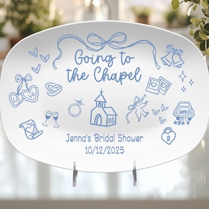 Op de afbeelding: Witte ovale serveerschaal met blauwe illustraties en tekst. De schaal toont de zin "Going to the Chapel" en diverse tekeningen met een bruiloftthema. De tekst "Jenna's Bridal Shower 10/12/2025" is ook opgenomen.
