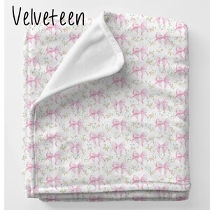 May include: A soft pink and white throw blanket with a repeating pattern of pink bows and small flowers. The blanket is folded in half and has a white fleece backing.