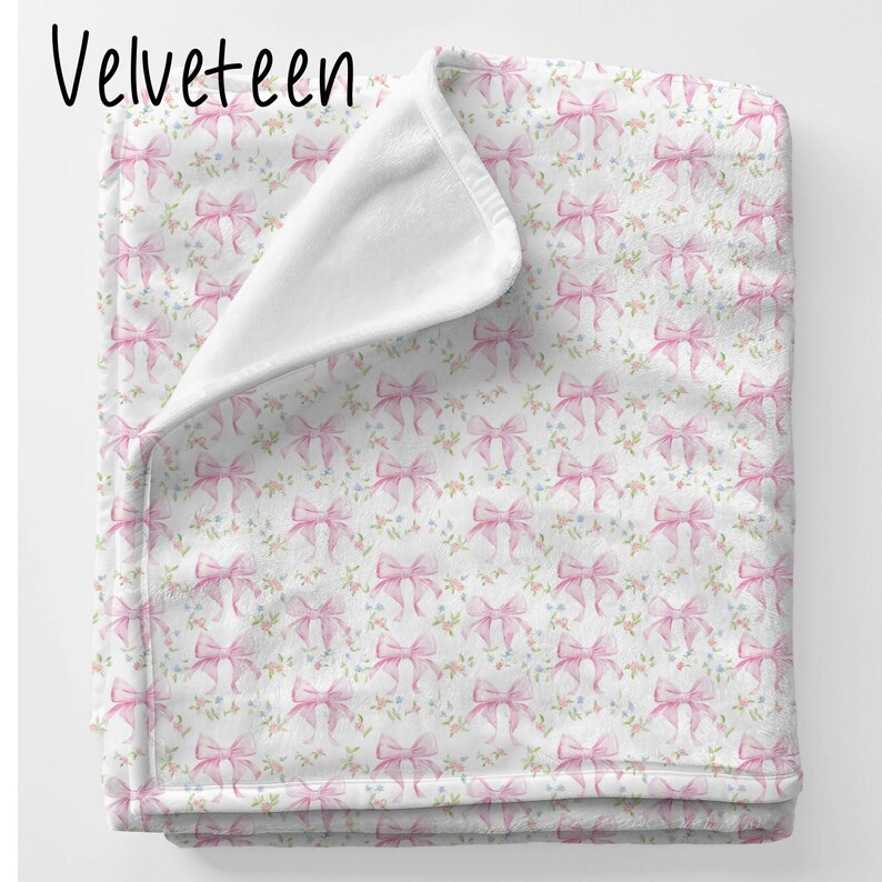 May include: A soft pink and white throw blanket with a repeating pattern of pink bows and small flowers. The blanket is folded in half and has a white fleece backing.
