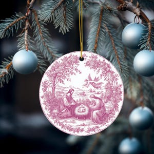 May include: A round Christmas ornament with a pink and white illustration of the Nativity scene. The ornament is hanging from a gold string against a backdrop of a Christmas tree and blue ornaments.