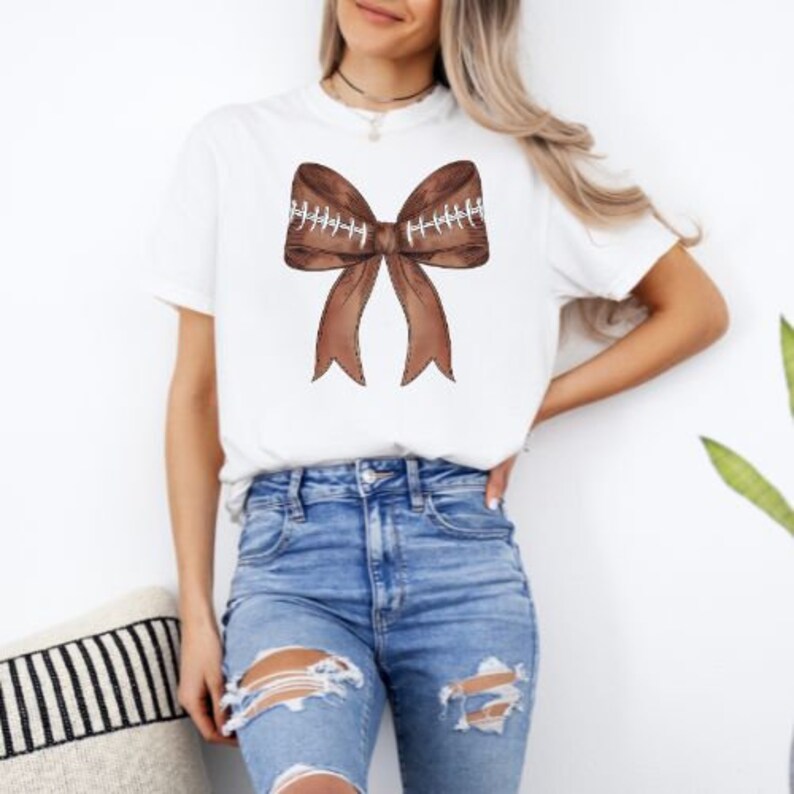 May include: A white t-shirt with a brown football-shaped bow on the front. The bow is made of brown fabric with white stitching.