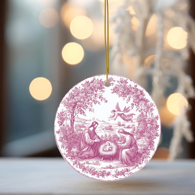 May include: Round Christmas ornament with a pink and white illustration of the Nativity scene. The ornament has a gold string for hanging. The illustration depicts Mary, Joseph, baby Jesus, and an angel in a pastoral setting.