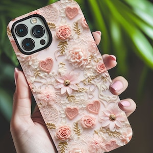 Puede incluir: Una funda de teléfono rosa y blanca con un diseño floral y corazones. La funda tiene un recorte para el objetivo de la cámara.