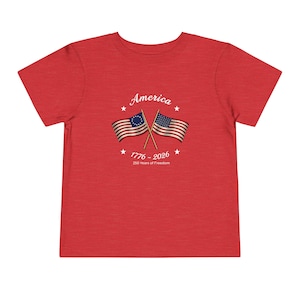 May include: Red t-shirt with the word "America" above two crossed American flags. The flags are from the Revolutionary War era. The text "1776 - 2026" and "250 Years of Freedom" are below the flags. The shirt is made of a soft material.