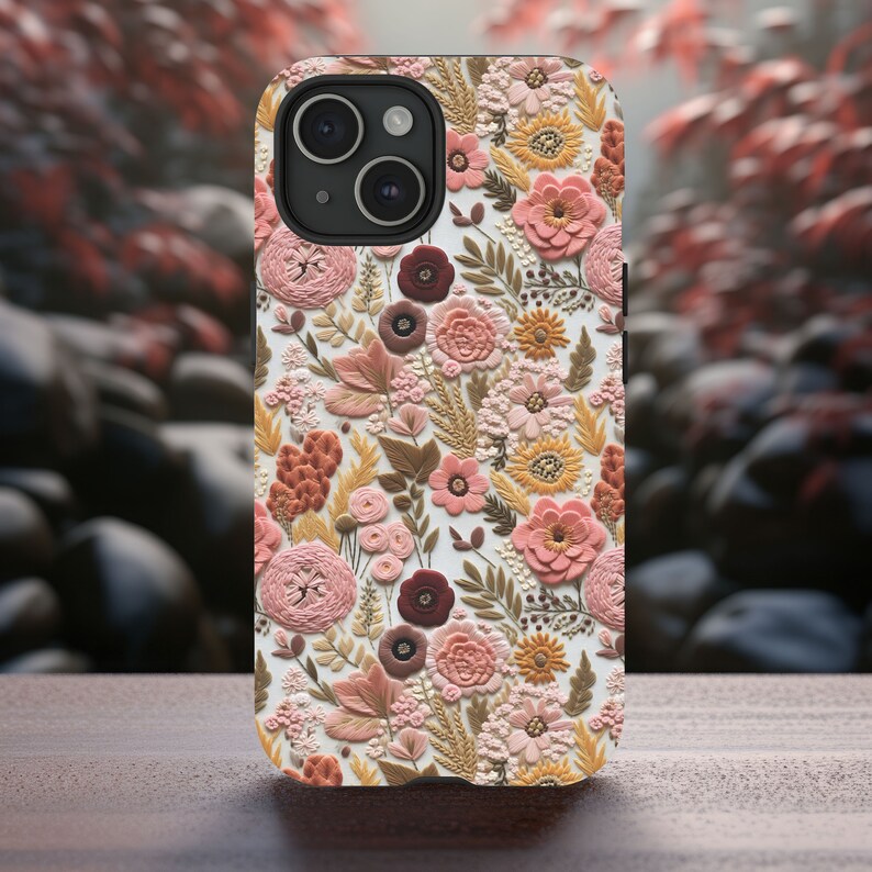 May include: A floral phone case featuring a detailed pattern of embossed flowers in shades of pink, peach, and brown. The case has a white background and a raised texture, giving it a 3D appearance. The phone case is on a wooden surface.