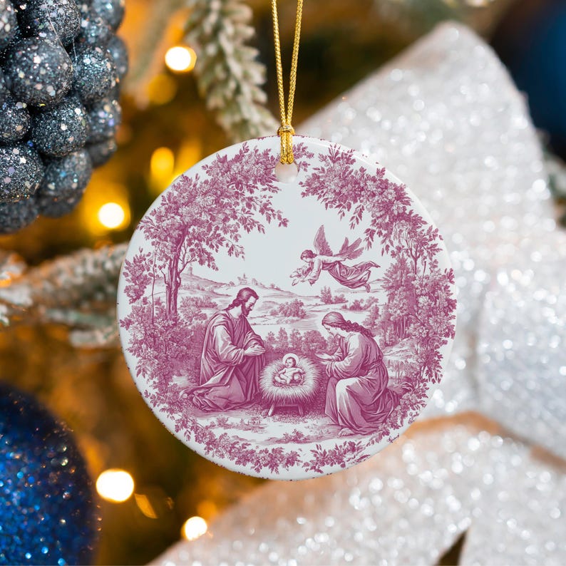May include: A round Christmas ornament with a religious scene in a toile design. The ornament features a burgundy and white color scheme, depicting the nativity scene with Jesus, Mary, and an angel. The ornament is hanging from a gold string.