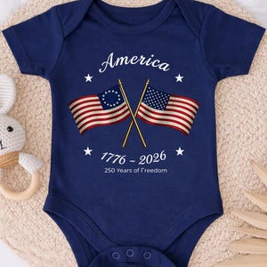 May include: Navy blue baby bodysuit with the word "America" and the dates "1776 - 2026" printed in white. The bodysuit features two crossed American flags and white stars. The text "250 Years of Freedom" is also printed on the bodysuit.