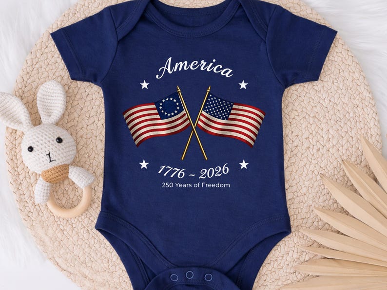 May include: Navy blue baby bodysuit with the word "America" and the dates "1776 - 2026" printed in white. The bodysuit features two crossed American flags and white stars. The text "250 Years of Freedom" is also printed on the bodysuit.