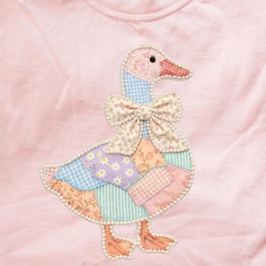 May include: A light pink long-sleeved shirt featuring a patchwork goose design. The goose has a pink head, a floral bow, and a body made of various patterned fabrics. The design is outlined with a pearl-like trim.