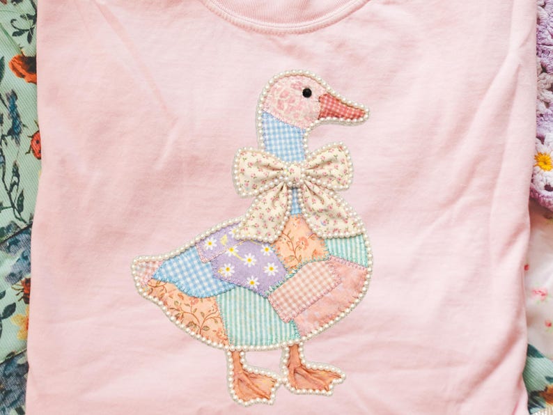 May include: A light pink long-sleeved shirt featuring a patchwork goose design. The goose has a pink head, a floral bow, and a body made of various patterned fabrics. The design is outlined with a pearl-like trim.
