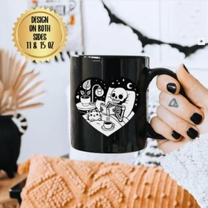 Puede incluir: Taza de café negra con un diseño blanco de un esqueleto en la cama leyendo un libro con un gato a su lado. El diseño tiene forma de corazón con estrellas y una luna creciente en el fondo. La taza está disponible en 11 o 15 onzas.