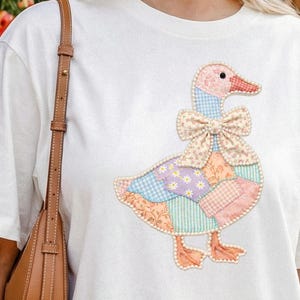 May include: White t-shirt featuring a colorful patchwork goose design. The goose has a pink head and feet, a patterned bow, and a body made of various pastel fabrics. The shirt is a casual, everyday wear item.