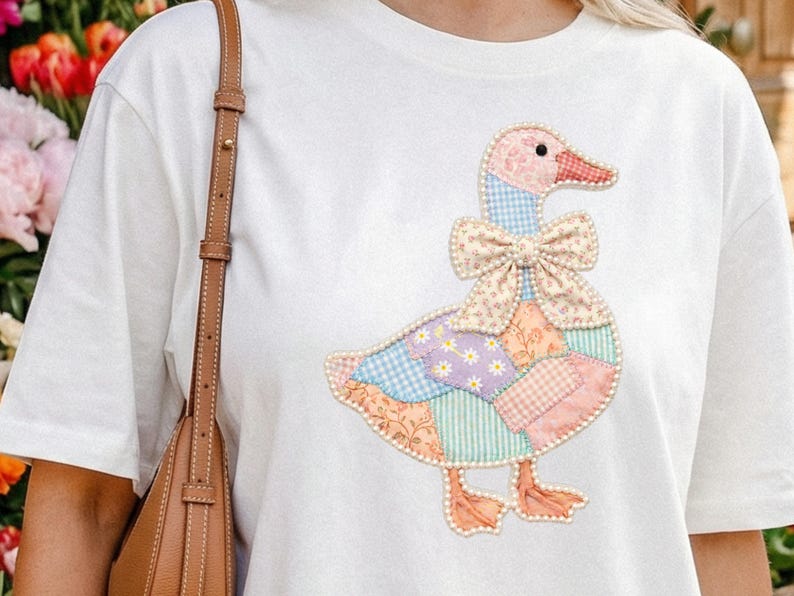 May include: White t-shirt featuring a colorful patchwork goose design. The goose has a pink head and feet, a patterned bow, and a body made of various pastel fabrics. The shirt is a casual, everyday wear item.