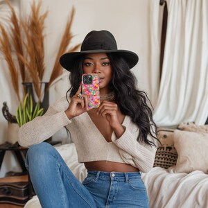 May include: A woman wearing a wide-brimmed black hat and a cream sweater is holding a colorful phone case in front of her face. The case has a floral pattern with pink, yellow, blue, and green colors.