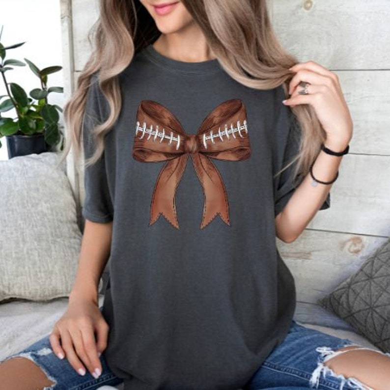 May include: A charcoal grey t-shirt with a brown football-themed bow on the front. The bow is made of two footballs tied together with a brown ribbon.