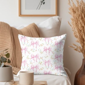 K&ouml;nnte beinhalten: Ein wei&szlig;es Kissen mit einer rosa Schleife und einem Blumenmuster. Das Kissen liegt auf einem wei&szlig;en Sofa mit einer braunen Decke und einer Vase mit Trockenblumen.