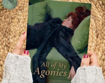 May include: A book cover featuring a painting of a woman in a black dress reclining on a green surface. The title "All of My Agonies" is printed in gold lettering. The image is held by a person with red nail polish.