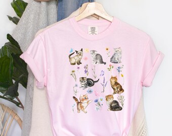 May include: A pink t-shirt with a graphic print of multiple kittens and flowers. The kittens are in various poses and colors, and the flowers are in shades of pink, purple, and yellow.
