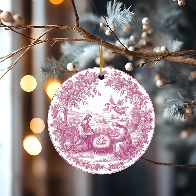 May include: A round Christmas ornament with a pink and white toile design depicting the Nativity scene. The ornament is hanging from a gold string and is surrounded by a blurred background of Christmas decorations.