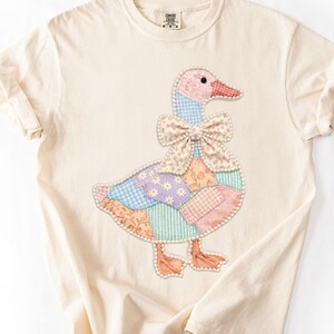 May include: Cream-colored t-shirt featuring a patchwork goose design. The goose has a pink head, a red beak, and a patterned bow. The body is made of various pastel-colored fabric patches. The shirt has rolled-up sleeves.