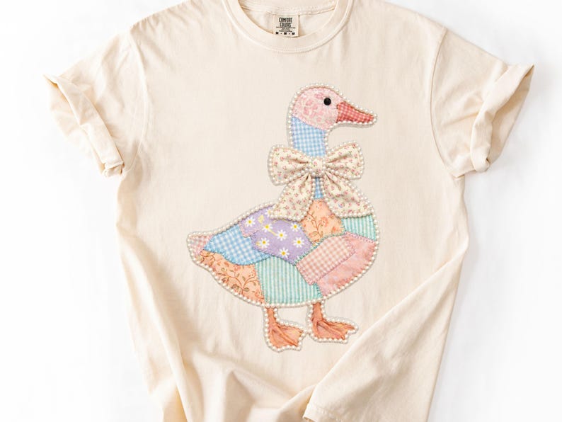 May include: Cream-colored t-shirt featuring a patchwork goose design. The goose has a pink head, a red beak, and a patterned bow. The body is made of various pastel-colored fabric patches. The shirt has rolled-up sleeves.