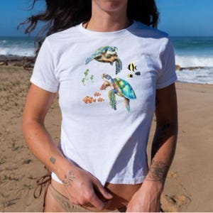 May include: White t-shirt with a colorful sea turtle design. The design features two sea turtles, coral, and fish.