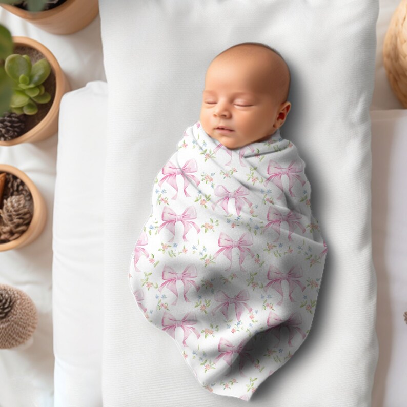 May include: A baby swaddled in a white swaddle blanket with a pink bow pattern. The baby is sleeping peacefully.