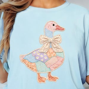 May include: Light blue t-shirt featuring a patchwork goose design. The goose has a pink head and feet, a bow tie, and a body made of various patterned fabrics in pastel colors. The shirt is a comfortable, casual style.