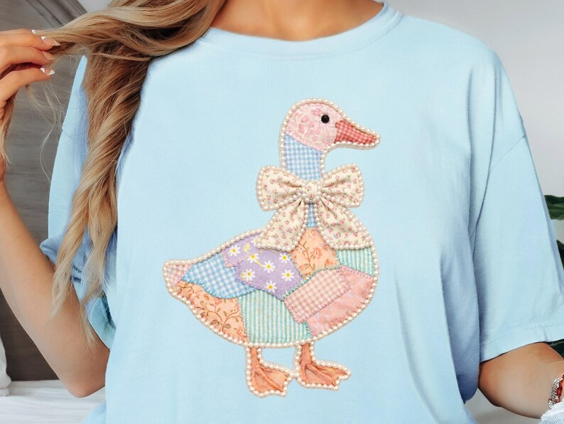 May include: Light blue t-shirt featuring a patchwork goose design. The goose has a pink head and feet, a bow tie, and a body made of various patterned fabrics in pastel colors. The shirt is a comfortable, casual style.