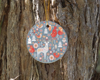 May include: A round, gray ceramic ornament with a Scandinavian folk art design. It features a white deer, red birds, and a fox, all surrounded by stylized trees and flowers.